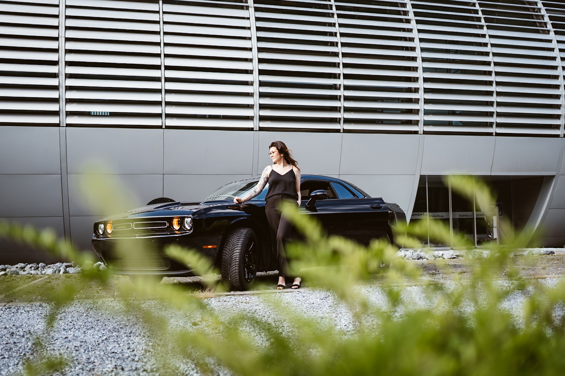 Julia im schwarzen Outfit vor ihrem Auto; breites Porträt mit grünen Zweigen im Vordergrund